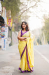 Woman in a yellow and purple saree standing on a street with trees in the background. www.laaki.co