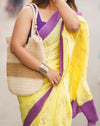 Woman wearing a yellow saree with purple accents, holding a woven bag. www.laaki.co
