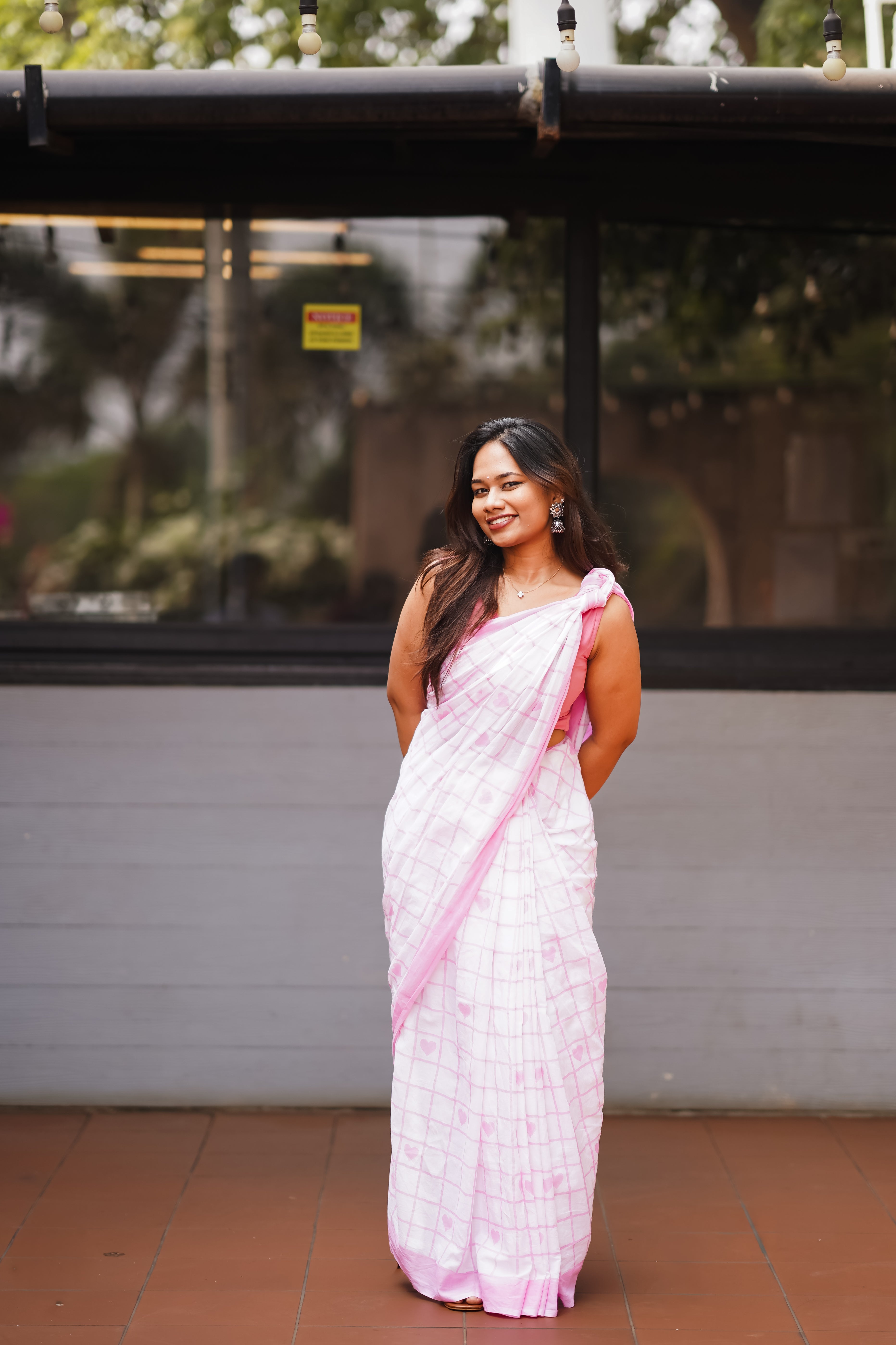 Woman in a white and pink saree standing in front of a building with reflective glass windows. laaki
