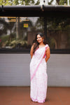 Woman in a white and pink saree standing in front of a building with reflective glass windows. laaki
