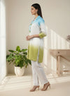 Woman in a light blue and green outfit standing in a bright room with a plant and table. www.laaki.co