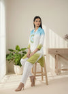 Woman sitting on a stool wearing a light blue and green outfit in a bright room with plants and a desk. www.laaki.co
