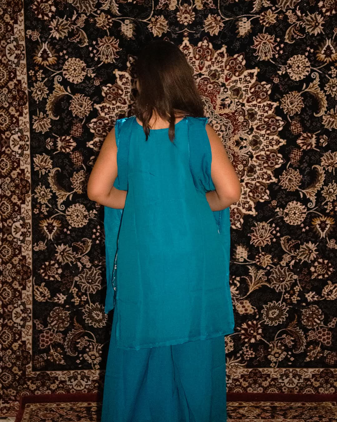 laaki Person wearing a teal outfit standing in front of a patterned rug