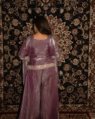 Person wearing a purple traditional outfit standing against a patterned wall. laaki