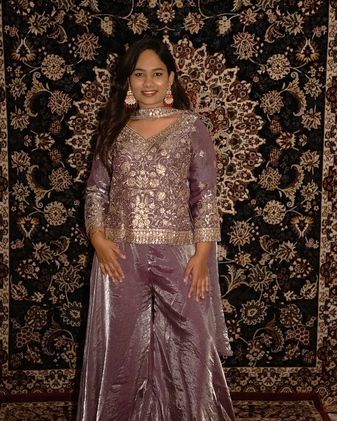 Woman in a purple traditional outfit standing against a patterned wall. laaki