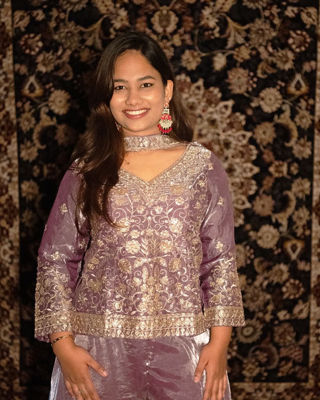 Woman wearing a purple traditional outfit with intricate patterns against a patterned wall. laaki