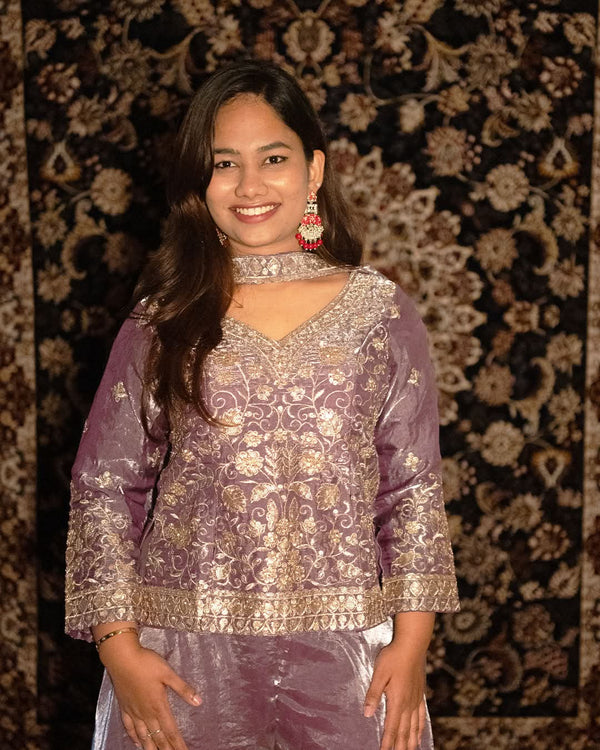 Woman wearing a purple traditional outfit with intricate patterns against a patterned wall. laaki