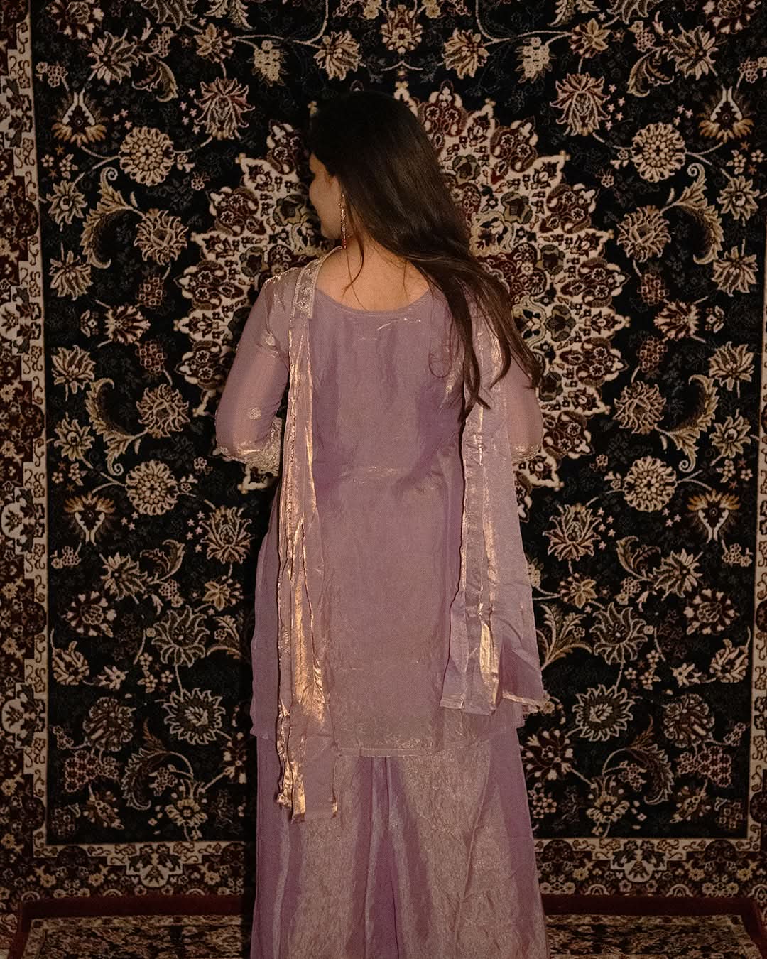 Woman in a lavender outfit standing against a patterned wall. sharas