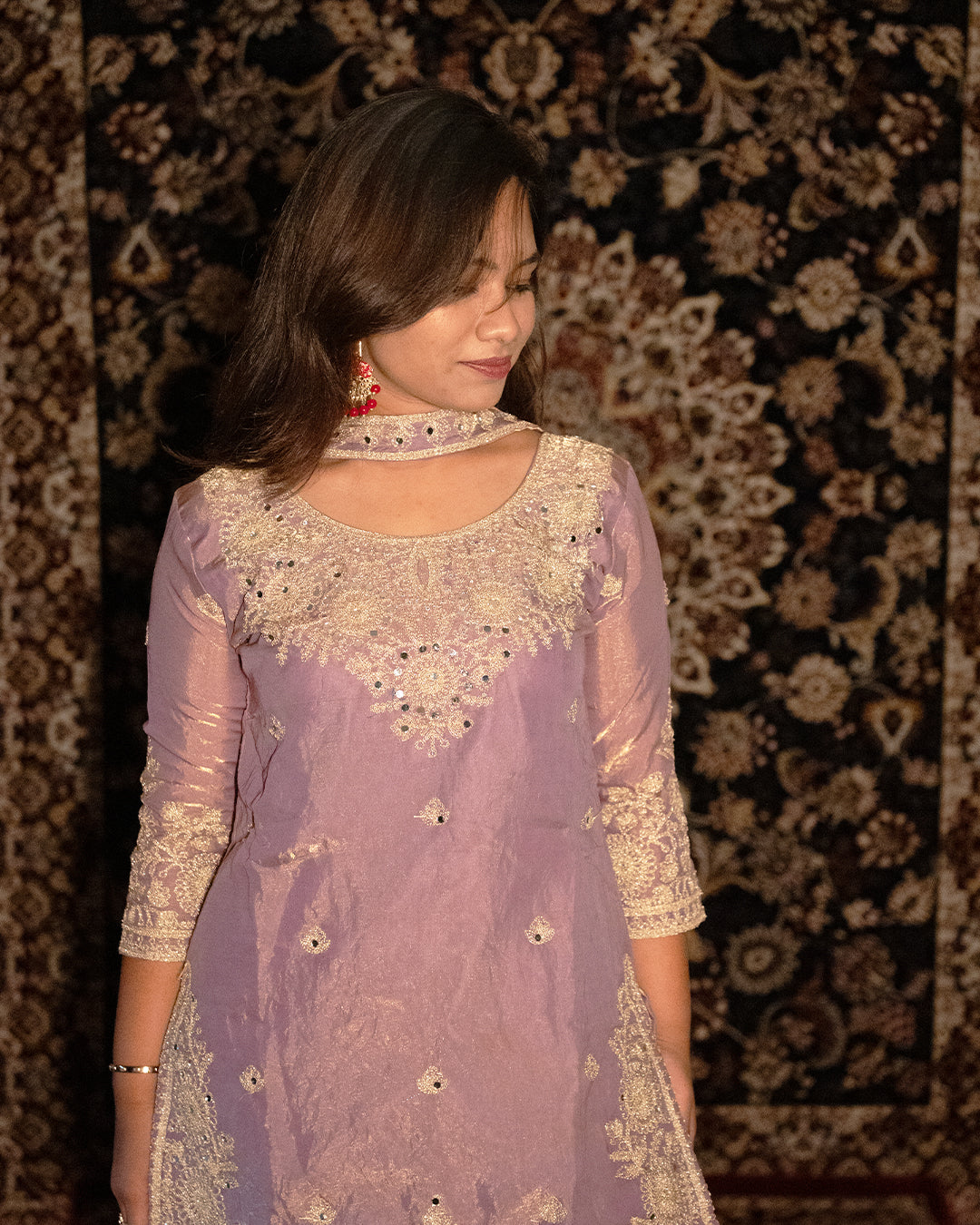 Woman wearing a lavender traditional outfit with intricate gold embroidery against a patterned rug background. www.laaki.co