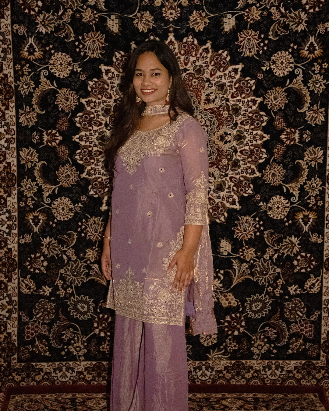 Woman in a lavender traditional outfit standing against an ornate patterned . www.laaki.co wall.