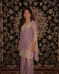 Woman in a lavender traditional outfit standing against an ornate patterned . www.laaki.co wall.