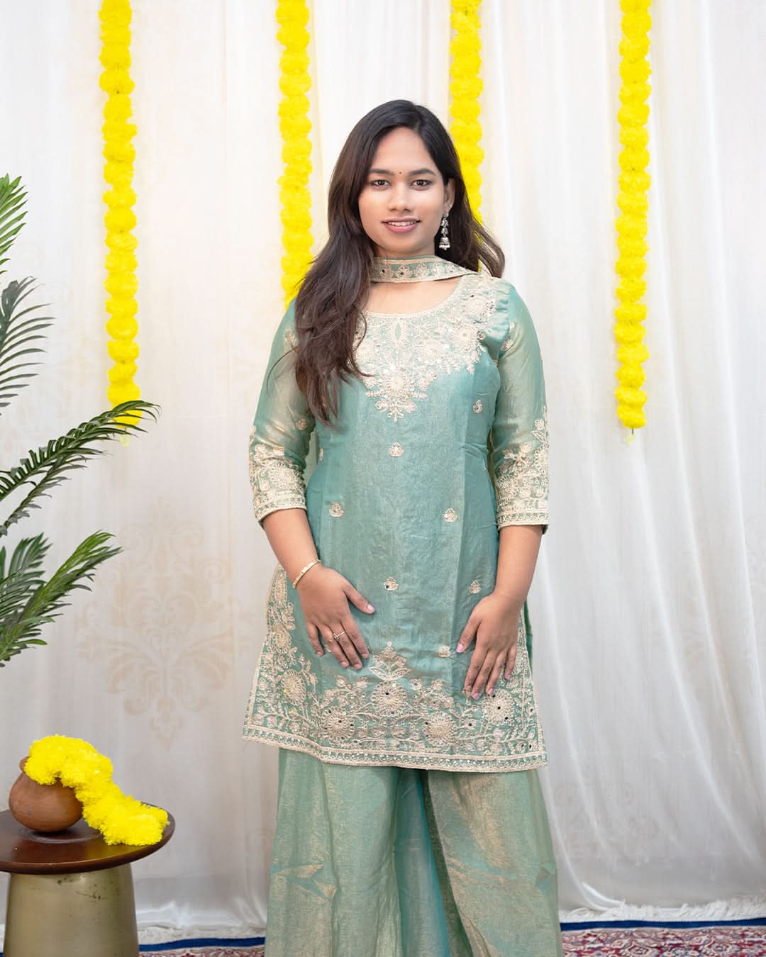 Woman in a light green traditional outfit standing against a white curtain with yellow decorative elements. laaki