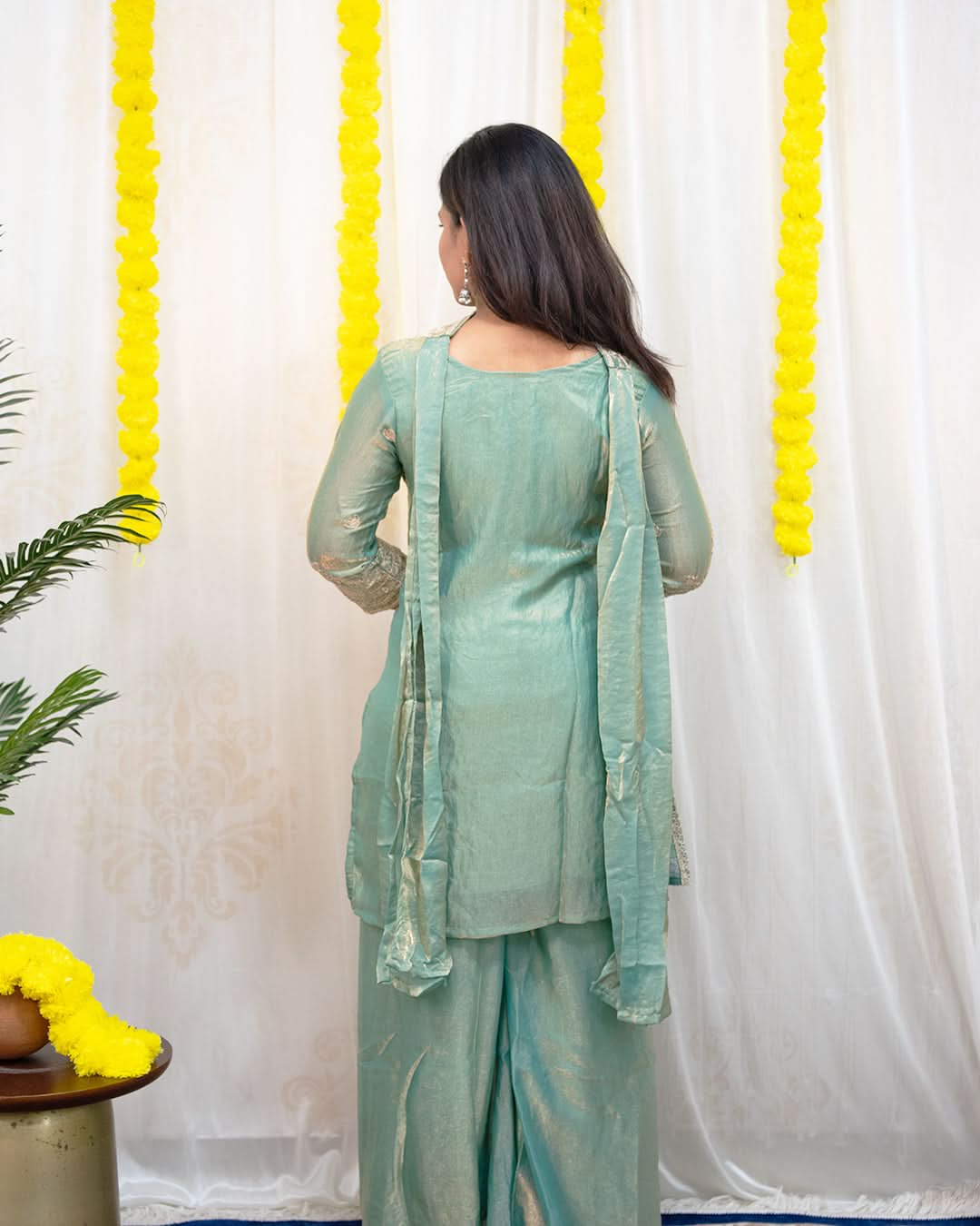 Woman in a light green saree standing against a white curtain with yellow decorative elements. laaki