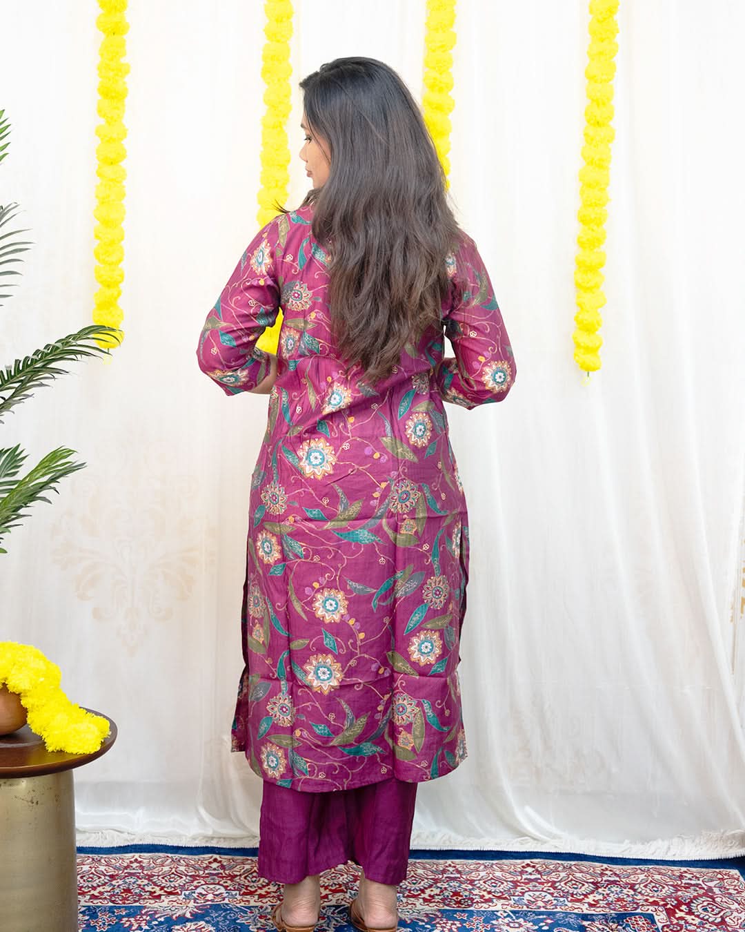 Woman in a pink floral kurta with purple pants standing against a white wall with yellow decorations. laaki