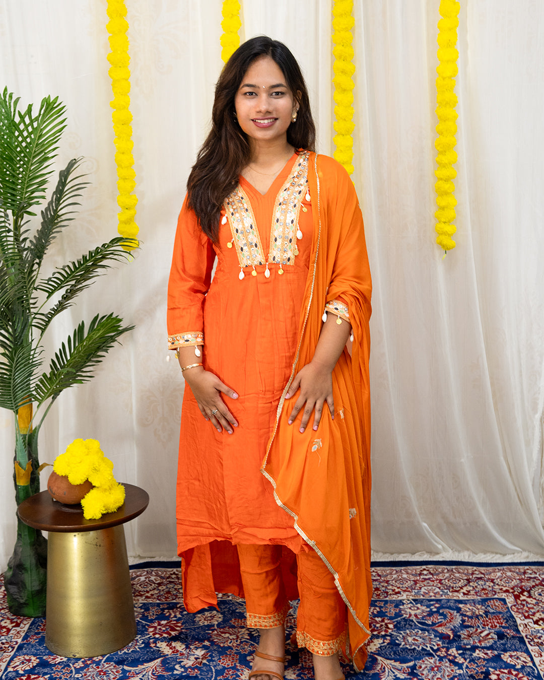 laaki Woman in an orange traditional outfit standing in a decorated room with yellow decorations and a plant.