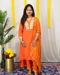 laaki Woman in an orange traditional outfit standing in a decorated room with yellow decorations and a plant.