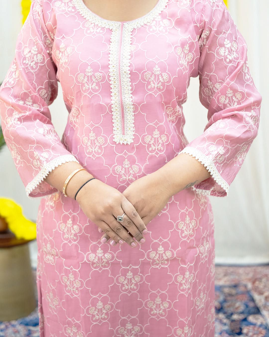 Person wearing a pink traditional outfit with white floral patterns indoors. laaki
