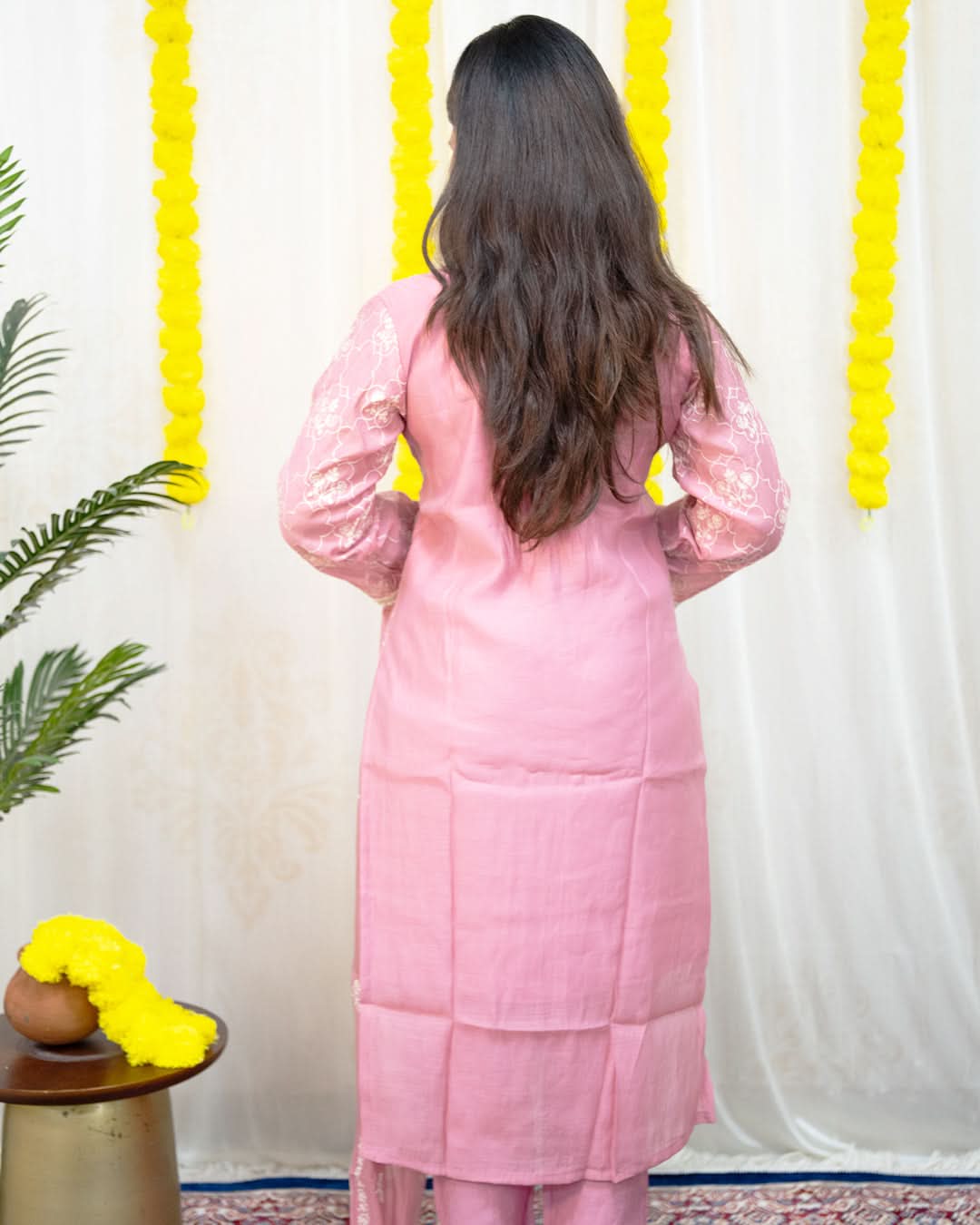 Person wearing a pink traditional outfit with a white curtain and yellow decorations in the background. laaki
