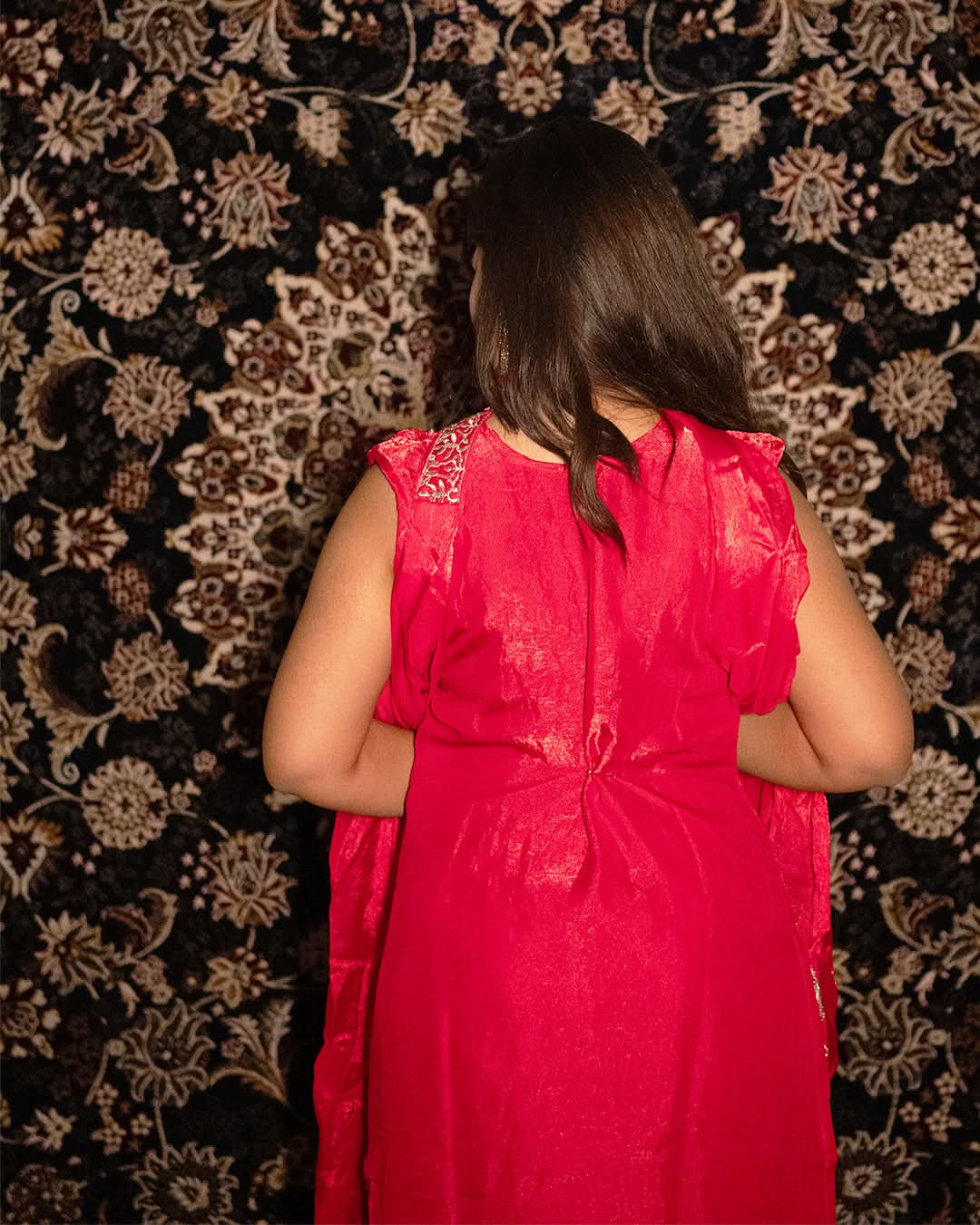 Person wearing a red dress with floral patterns against a patterned wall. laaki