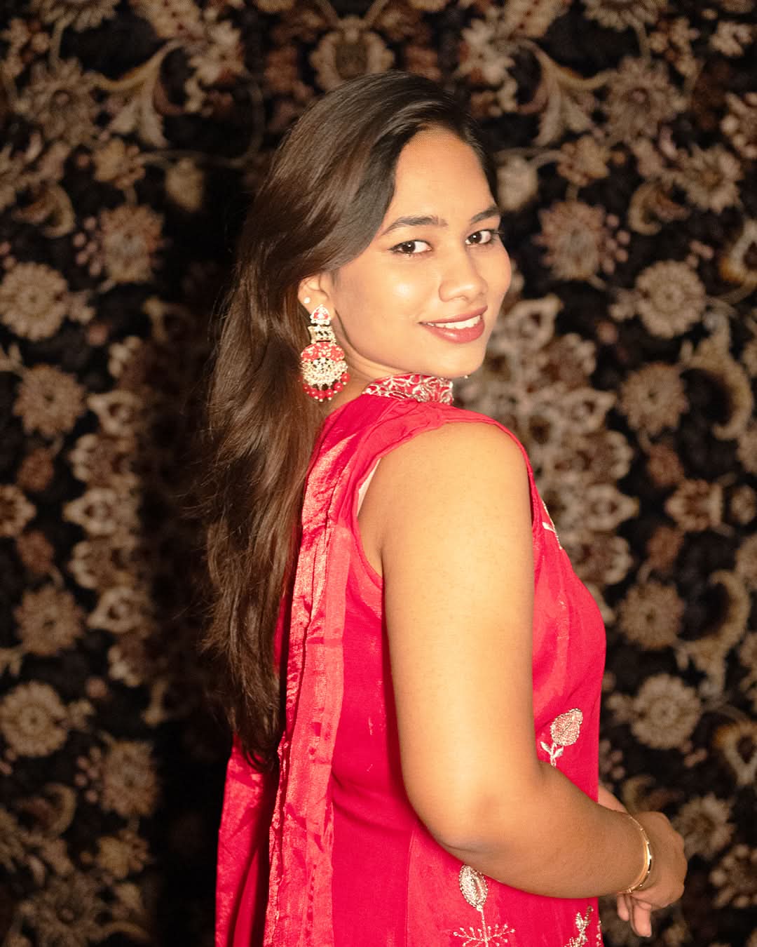 Woman in a red traditional outfit with floral patterns against a decorative laaki background