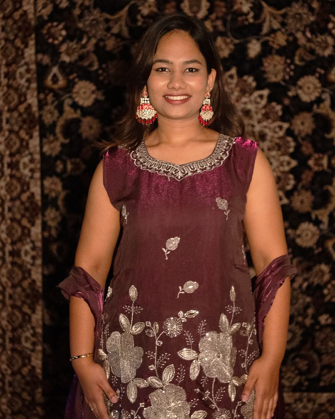 Woman wearing a maroon traditional outfit with floral patterns and silver embellishments against a patterned background. laaki