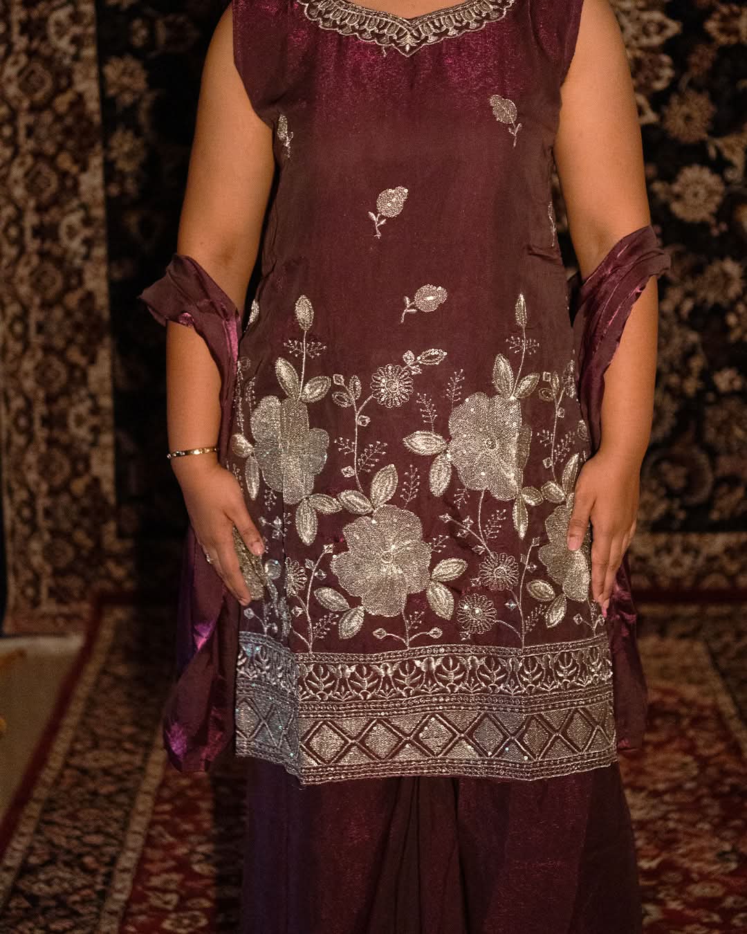Maroon traditional outfit with floral embroidery on a patterned carpet background. laai