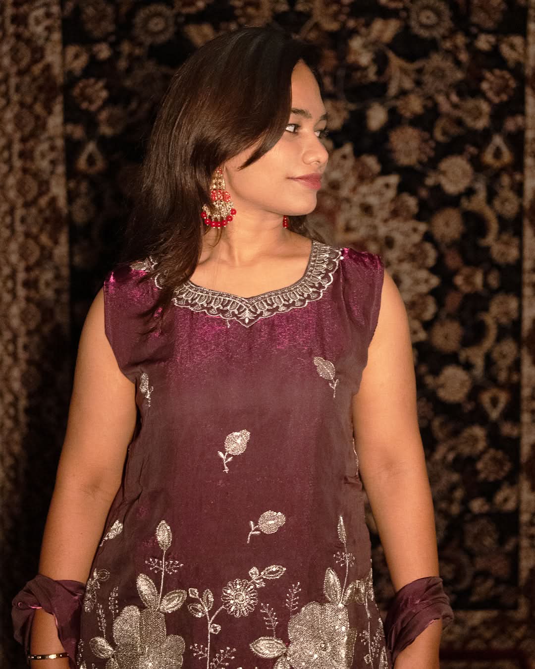 Woman wearing a maroon traditional outfit with silver embroidery against a patterned background laaki.