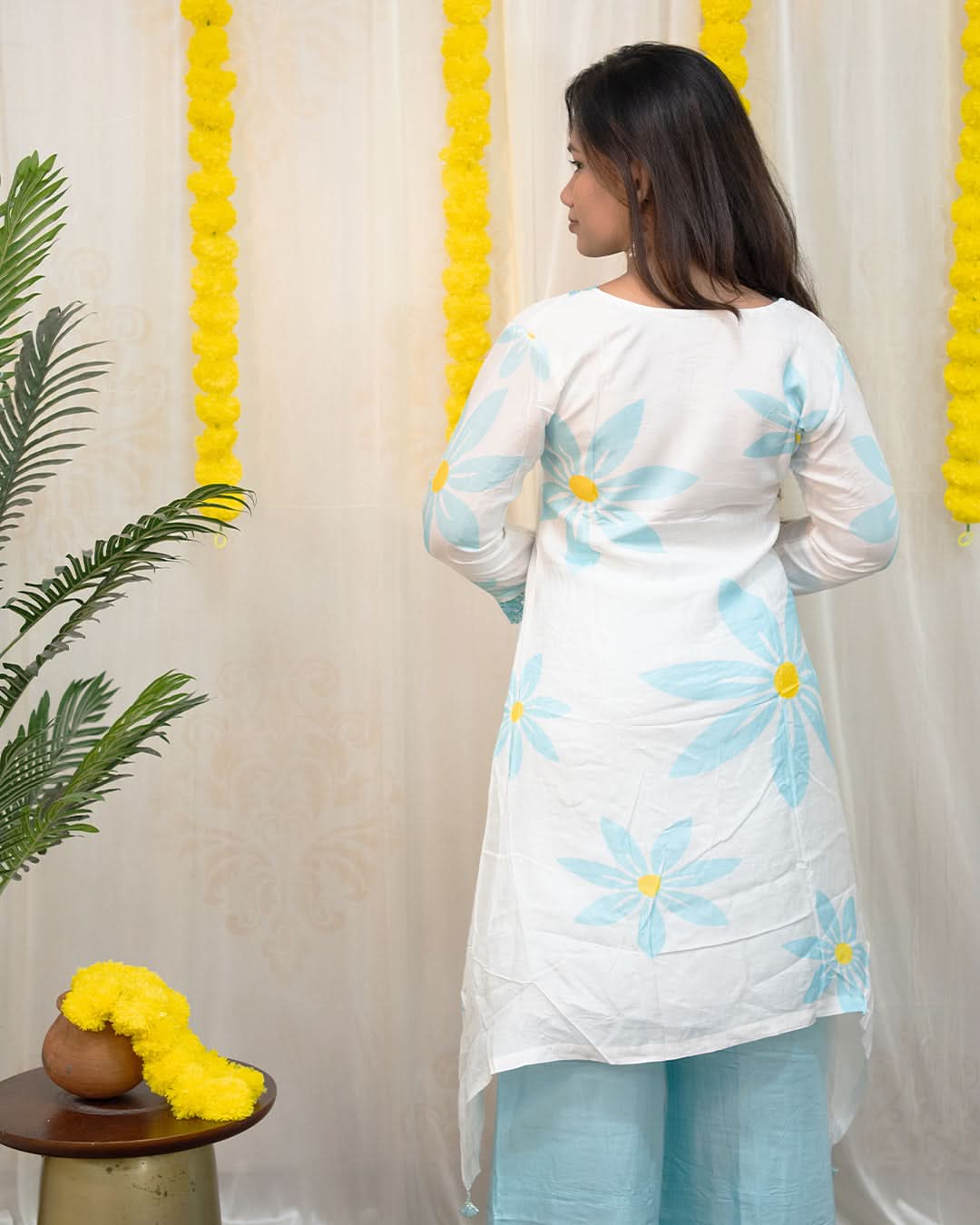 Woman wearing a white kurta with blue floral patterns against a decorative curtain background. www.laaki.co