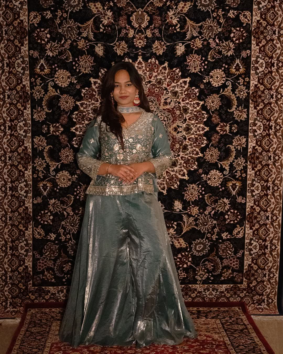Woman in a silver traditional outfit standing against a patterned wall. laaki