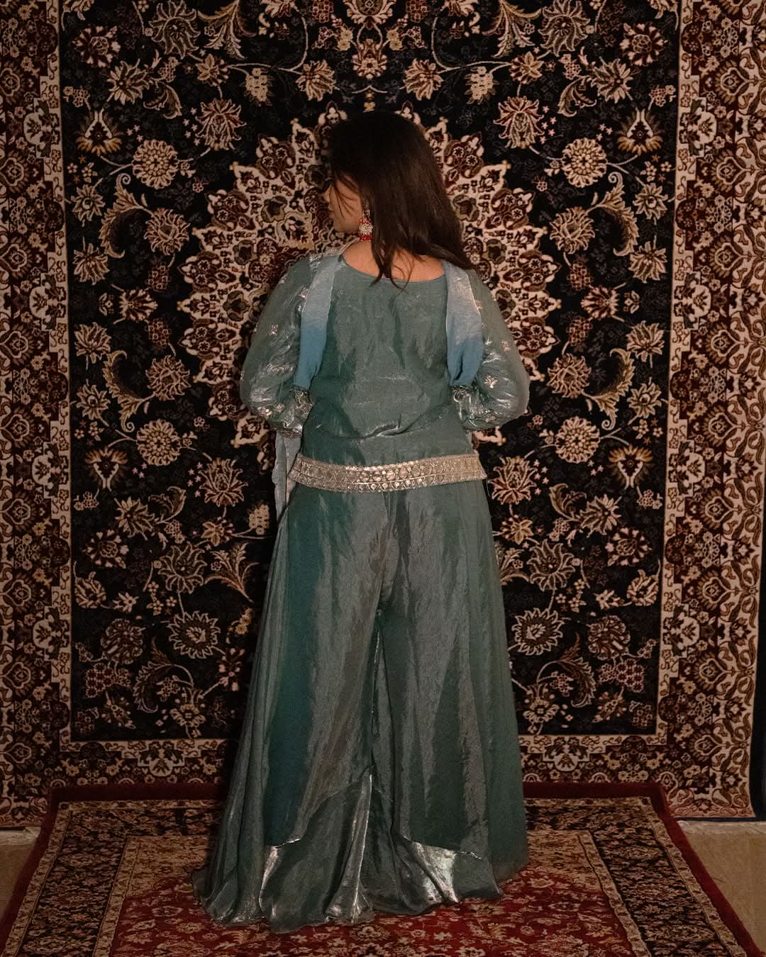 Person wearing a teal outfit with a patterned rug in the background. laaki