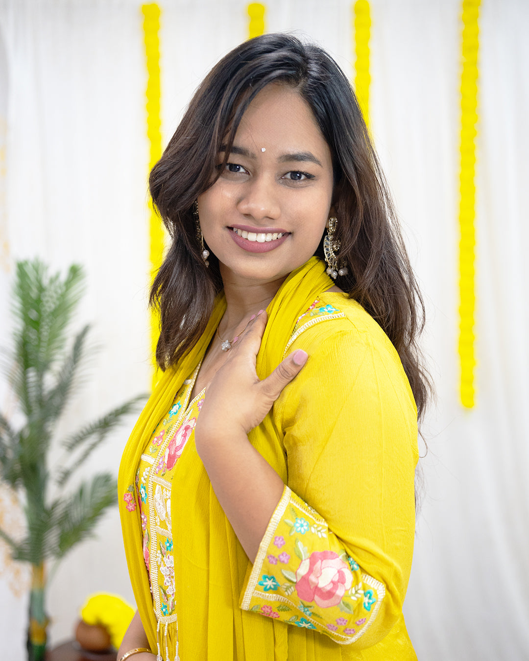 laaki Woman wearing a yellow traditional outfit with floral embroidery, smiling in front of a white wall with yellow vertical stripes.